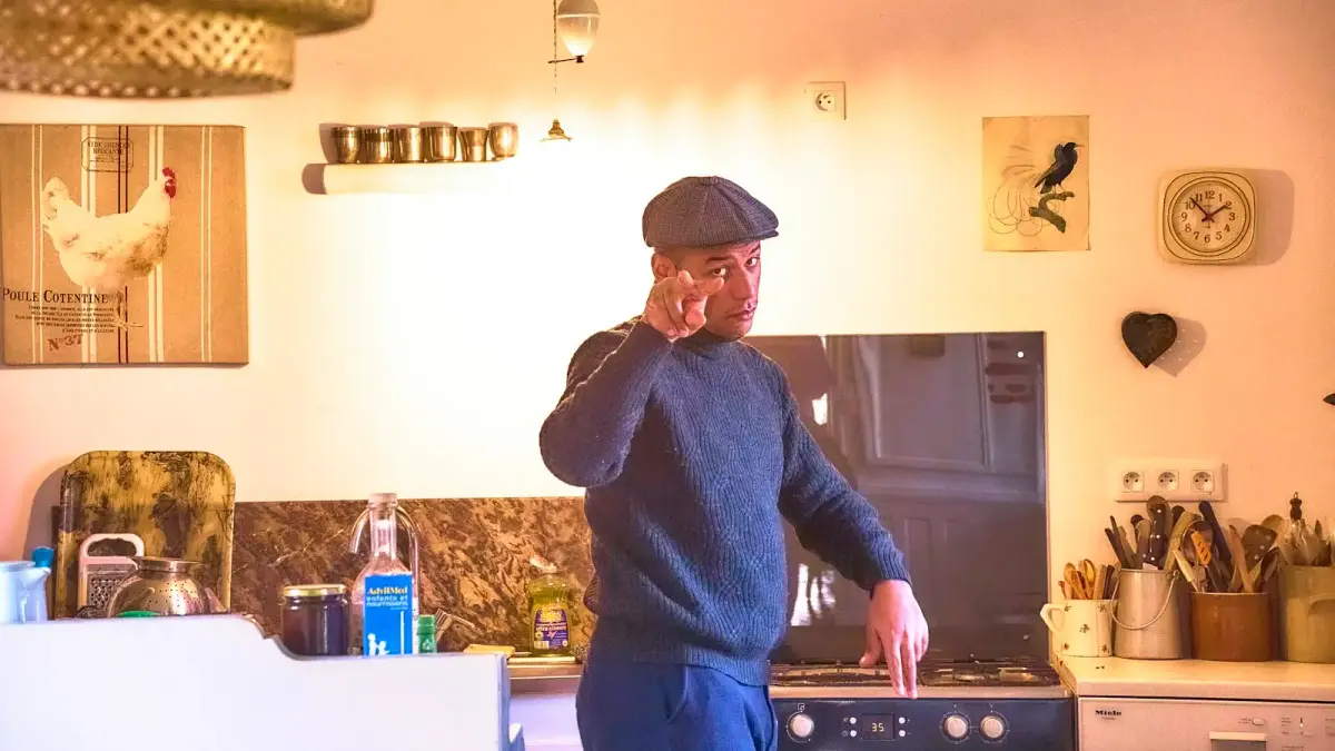 A man standing in a kitchen holding a phone, hesitating before dialing